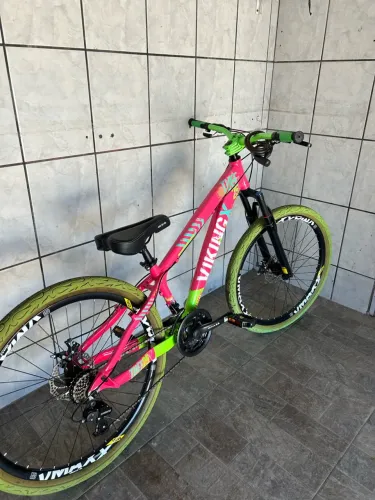 Bicicleta do tipo Mountain Bike (MTB), com visual esportivo e cores neon (rosa e verde).