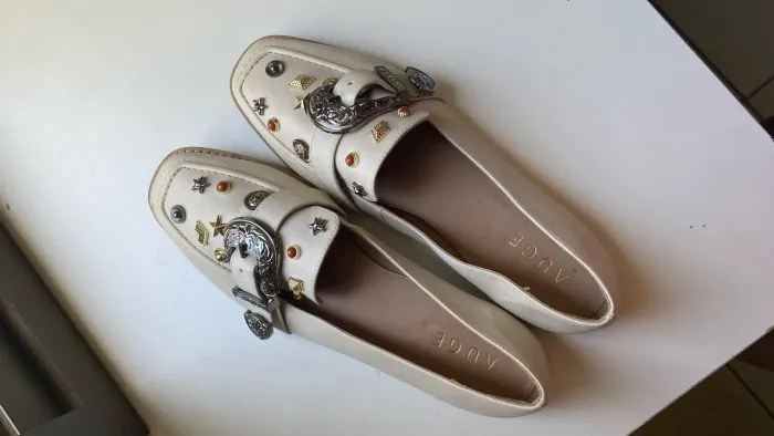 Mocassins off-white da marca Auge, decorados com tachas metálicas e fivelas estilo western
